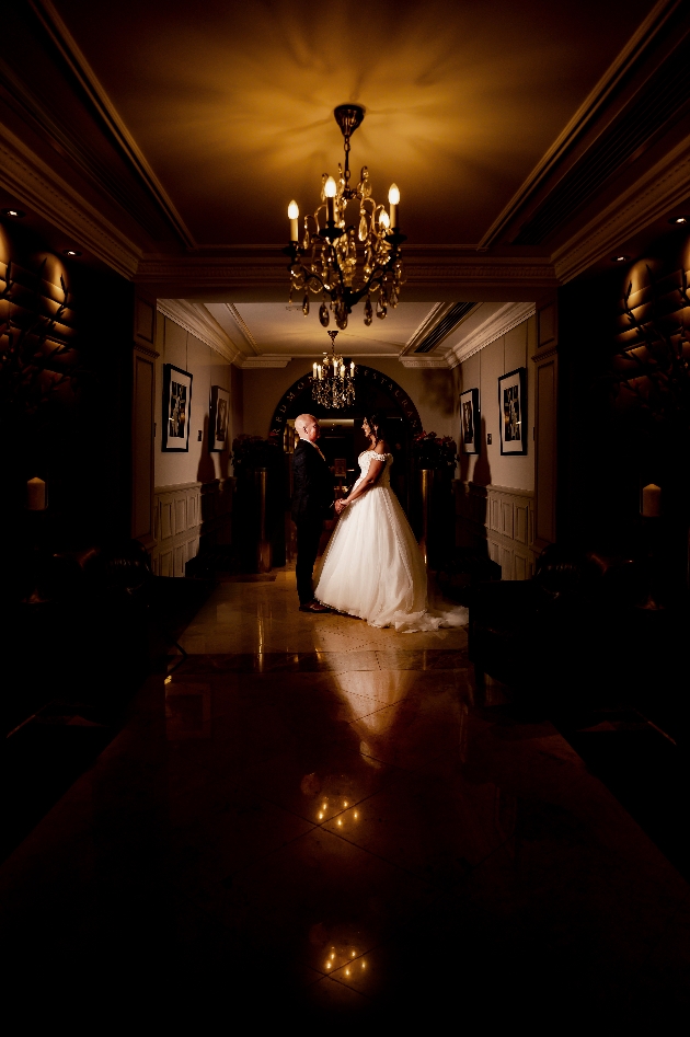couple holding hands facing each other, at the end of a long hallway with chandeliers