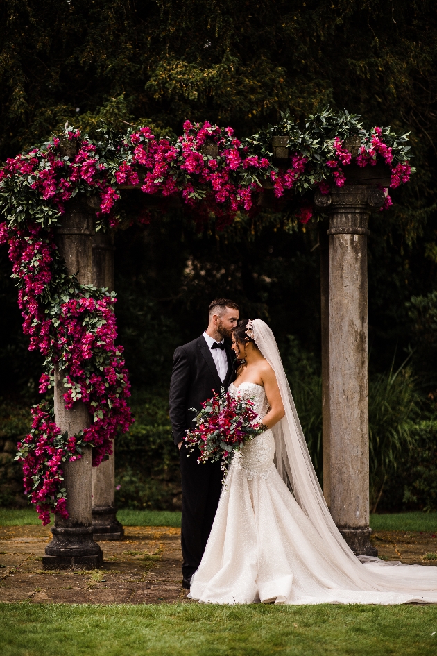 Bride and groom standing beneath a stone pagoda at Eaves Hall covered in bougainvillea