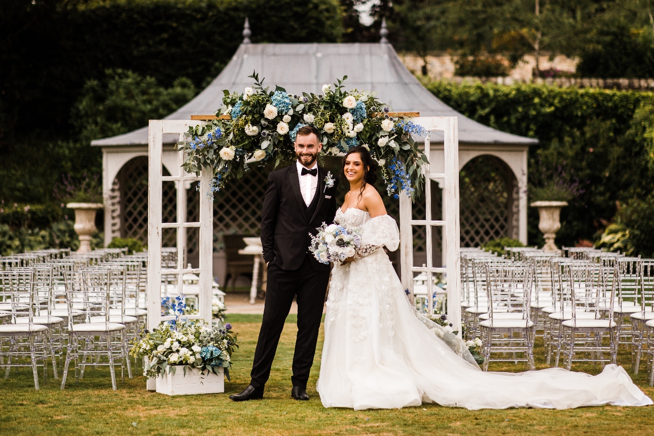 Couple in front of ceremony area at Eaves Hall with upcycled door arch and blue and white flowers