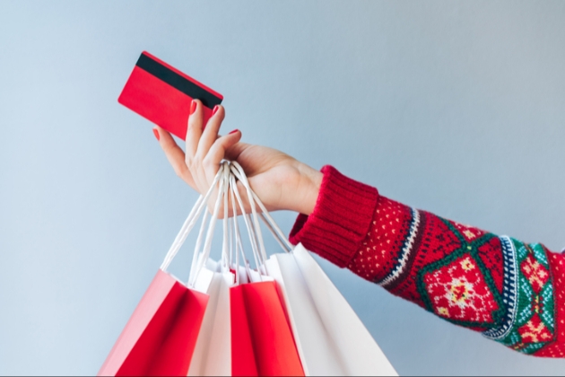 The arm of a woman wearing a christmas jumper holding out her credit card with christmas shopping bags on her arm 