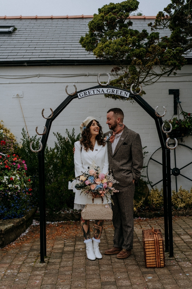 Eloping couple with luggage standing beneath Gretna Green sign