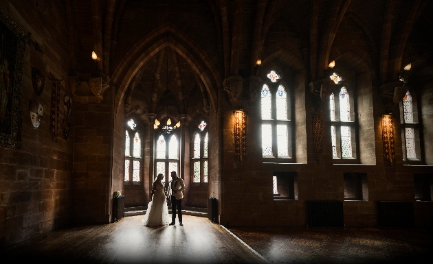 Bride and groom lit by daylight from dramatic gothic windows in castle