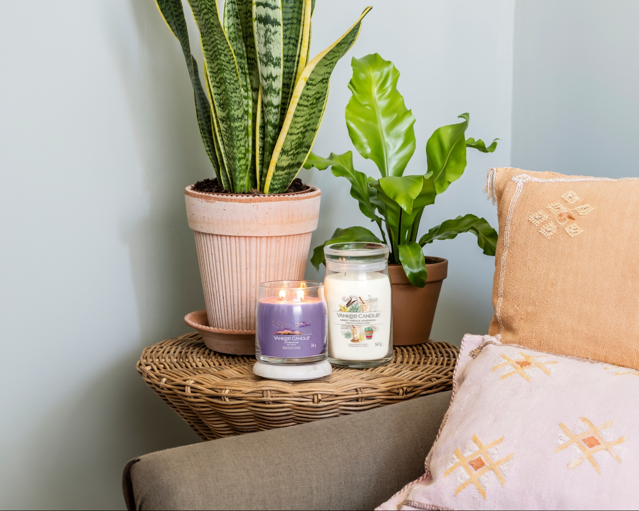 two candles on a rattan table with plant on it next to chair