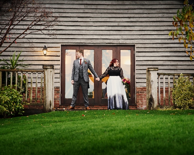 bride and groom and in hand looking away from each other in front of a wooden cabin, bride's dress is black with white and grey skirt