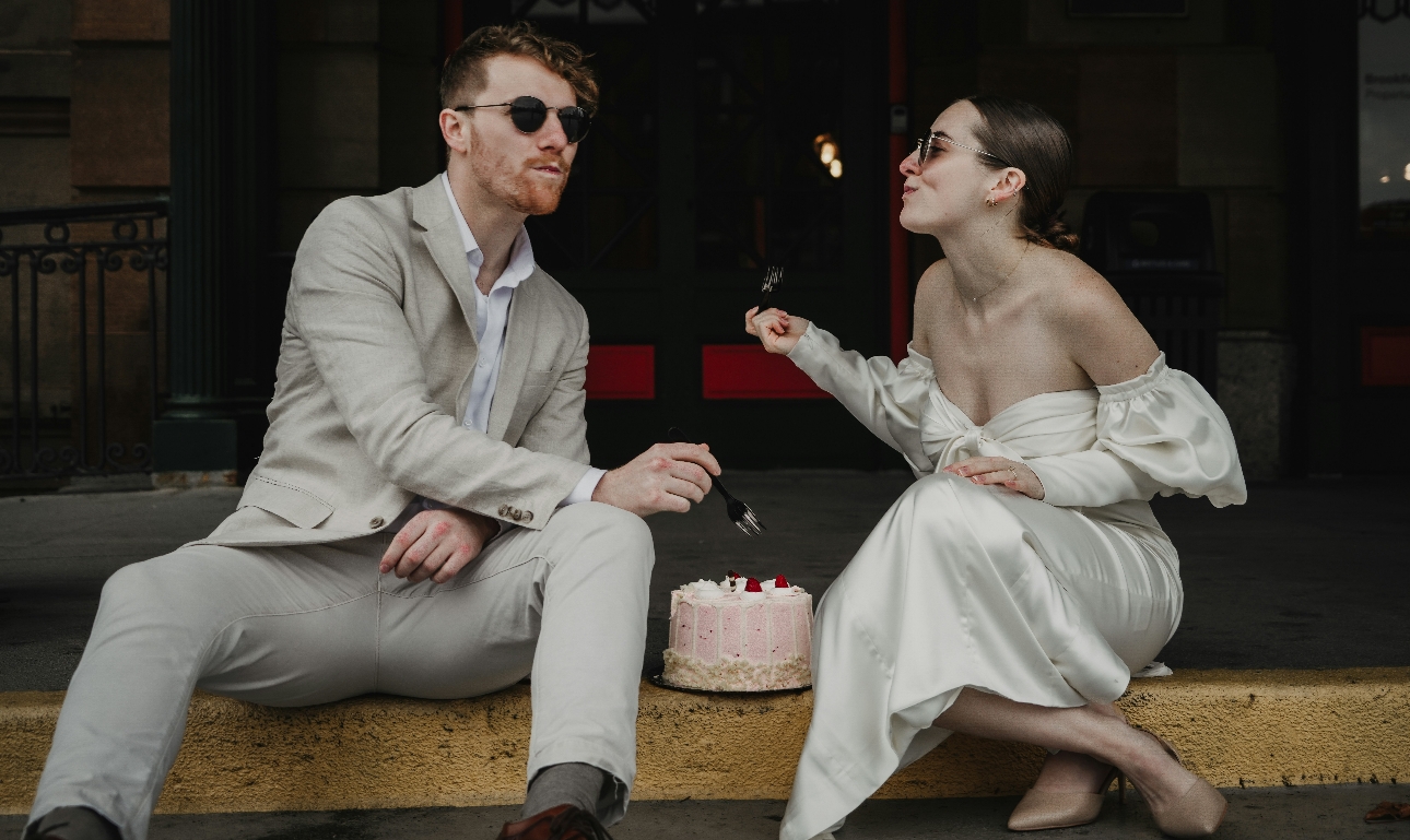 bride and groom sitting on the curb eating wedding cake